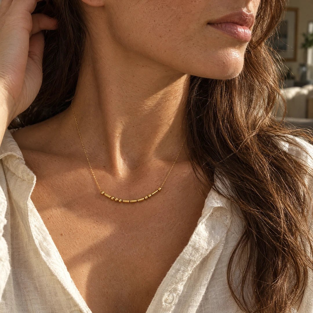 A close-up lifestyle shot of a woman wearing a delicate gold necklace with a curved row of small tube beads. She is wearing a light beige linen shirt, and the background is softly blurred with warm sunlight highlighting her skin and hair.